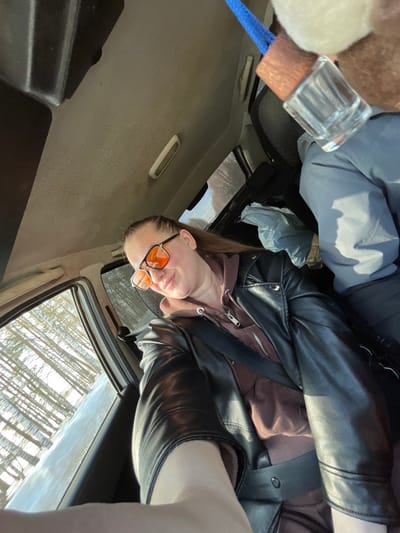 An image depicts the interior of a vehicle within the city of Nekrasovo, Russia. An adult female is positioned in the foreground, seated in what appears to be the front passenger seat. She has light brown hair, pulled back, and wears orange-tinted aviator-style sunglasses. Her expression is a slight smile directed towards the camera. She is dressed in a black leather-style jacket over a brown hooded sweatshirt and is secured by a black seatbelt across her chest and lap. Her left arm is extended towards the camera, suggesting she is taking a selfie.
To her right and slightly behind, the torso and legs of a second individual, wearing a grey or light blue jacket, are visible, likely occupying the driver's seat or another rear passenger seat. A light blue plastic bag is visible on a seat or floor in the mid-ground. Hanging from the overhead area of the vehicle, a clear glass bottle with a wooden top and a blue cord is partially visible. The vehicle's headliner is light grey and shows signs of wear.
Through the windows, a bright, sunny exterior environment is visible. This includes a dense collection of slender, light-colored tree trunks, indicative of a forest, possibly a birch grove, with what appears to be snow or frost on the ground, suggesting a winter or early spring season. The overall lighting is bright and natural.