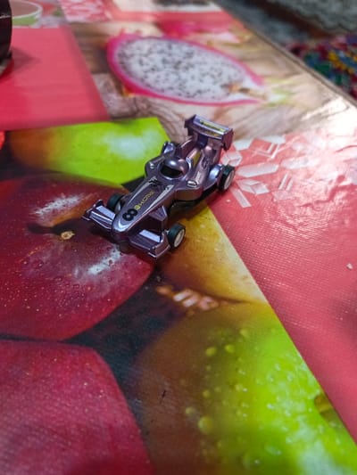 A miniature purple Formula 1 style race car, displaying the number '8' and the text "HCMG" on its side, and "JAGGER" on its rear wing, rests on a patterned surface. The surface, likely a tablecloth or placemat, features a vibrant print of various fruits, including red apples, a green fruit resembling a lime or green apple, and a cross-section of a dragon fruit. Water droplets are visible on the printed fruit images. The scene is located in the city of Akaltyn, Uzbekistan.