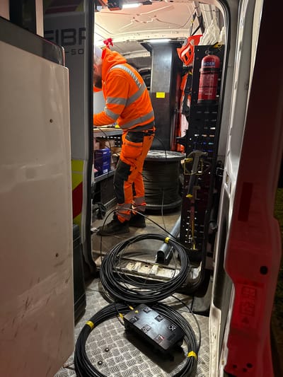 A man wearing an orange high-visibility jacket is bent over, working inside the rear of a van. The interior of the van is equipped with storage racks holding equipment, including a fire extinguisher. A large spool of cable and coils of black fiber optic cable are on the floor of the van. The van appears to be a utility vehicle associated with fiber optic installation, based on the "FIBRE" logo on the side. The location is potentially within Mid Suffolk, United Kingdom.