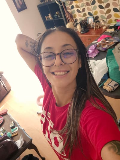 The image shows a woman with long, dark hair and glasses, wearing a red t-shirt, taking a selfie. She has a nose ring and tattoos on her arm. She is leaning back with her arm behind her head, smiling at the camera. The background appears to be a room with shelves filled with figurines and various items on a dresser. There is also a table in the foreground with several objects on it, including a wallet, cigarettes, and a can. The lighting suggests it is daytime, with a bright light source coming from the left side of the frame. The overall scene is casual and appears to be taken indoors, possibly in a home environment. The question about the location in Bridlington, United Kingdom is not answerable from the visual information provided in the image.