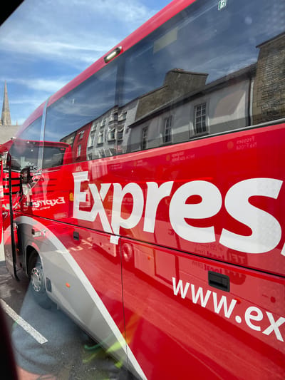 The image shows the side of a bright red tour bus with white lettering that reads "Express". The bus is parked on asphalt with white lines visible on the ground. In the reflection on the bus windows, buildings with traditional architecture are visible, including a church spire on the left. The sky above is blue with scattered clouds. The bus has silver accents along its lower body and a silver stripe. The context of Donegal, Ireland is not evident from the image itself but is provided as additional information.