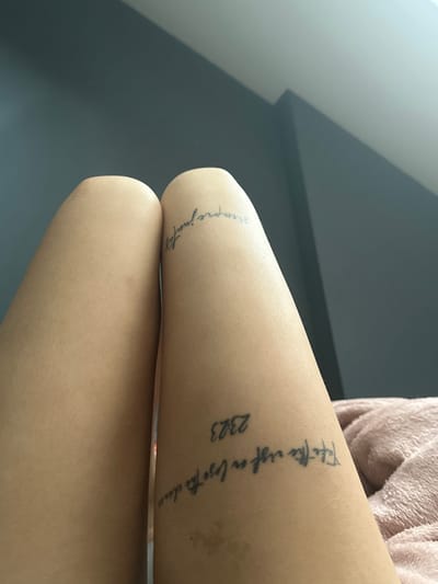 The image shows a close-up view of a person's legs, with tattoos on the inner thighs. On the left leg, there appears to be a tattoo on the upper thigh, but it is not clearly legible. On the right leg, there are two visible tattoos. The upper tattoo reads "siempre juntos" in cursive. Below this, another tattoo reads "2323" followed by the phrase "take the risk or lose the chance" in cursive. The person is lying down on a pink, fuzzy blanket, with a grey wall in the background. The lighting suggests an indoor setting, possibly a bedroom. There is no indication of the location being in Heerhugowaard, Netherlands.