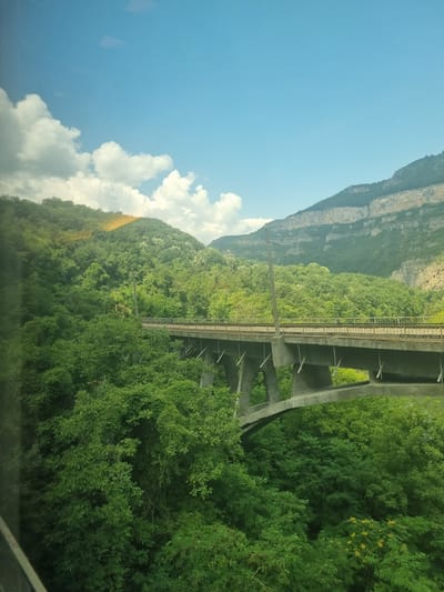 A concrete railway bridge spans a lush, tree-covered gorge under a bright blue sky with scattered white clouds. The bridge's piers are visible supporting the structure, which has two sets of parallel railway tracks running across it. Dense green foliage covers the slopes of the gorge, with the trees reaching up towards the bridge. In the background, a steep, rocky mountain face is visible, with some vegetation clinging to its sides. The scene suggests a rural or natural setting, likely within the environs of Milanovo, Bulgaria, known for its picturesque landscapes and railway infrastructure. No people or specific actions are depicted in the image.