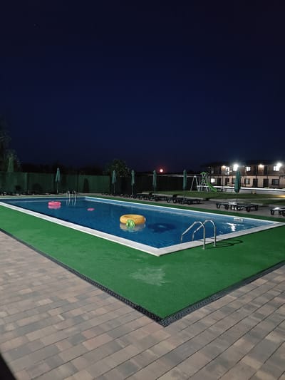 The image depicts an outdoor swimming pool area at night in Shkurupiivka, Ukraine. The pool is rectangular and filled with blue water, illuminated by overhead lights and the reflection of building lights. Two pink inflatable rings and one yellow inflatable ring are floating in the pool. A metal ladder is visible on the edge of the pool. The pool is surrounded by green artificial turf, which then transitions to a paved walkway made of interlocking bricks in shades of brown and grey. In the background, there are several lounge chairs and green umbrellas. Beyond the pool area, a two-story building with illuminated windows is visible. A children's playground with a slide is also present. The sky is dark blue, indicating nighttime, and a faint red circular object, possibly the moon or a distant light source, is visible in the upper left portion of the sky. The overall scene suggests a recreational facility at dusk or night.