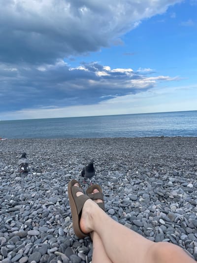 The image depicts a person's legs and feet, wearing brown sandals, crossed and resting on a pebble beach. Two pigeons are visible on the beach, one closer to the viewer and another further away. The sea stretches out beyond the beach, meeting a cloudy sky. The scene is set on a beach in the Livadiyskyi settlement council, Ukraine.