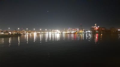 This image depicts a night view of the waterfront in Dock Sud, Argentina, characterized by a dark sky and illuminated city structures reflected on the rippling water. The foreground is dominated by a body of dark water, with scattered reflections of lights breaking the surface. In the midground, a bridge spans across the scene, with rows of lights illuminating its structure and the surrounding area. Beyond the bridge, a cityscape is visible, featuring various buildings, some of which are brightly lit with colorful lights, including red and white. A crane is also visible near the waterfront, suggesting industrial activity. The overall impression is a bustling urban environment at night, with the water serving as a reflective canvas for the city's glow.