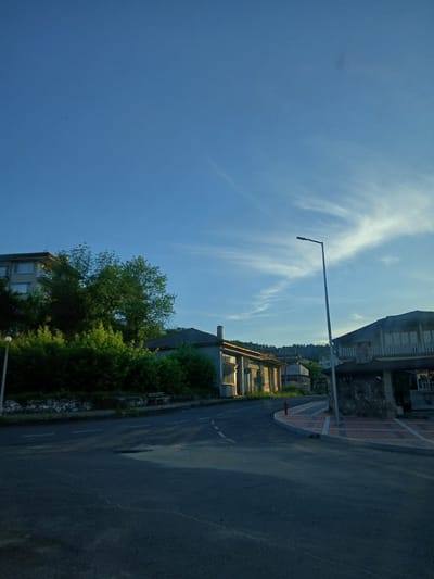 The image depicts a street scene in the city of Byal Izvor, Bulgaria, under a clear blue sky with wispy clouds. In the foreground, an asphalt road curves to the left, marked with white dashed lines. To the right of the road, a paved walkway with a brick pattern leads past a building with a stone-faced entrance. A red fire hydrant is visible near the corner of the walkway. A tall, slender lamppost stands on the right side of the road.

In the background, a long, low-rise building with multiple windows and a gray roof is situated on the left side of the street. Behind this building and extending further to the left, lush green trees and bushes create a dense natural barrier. A multi-story apartment building with visible windows is visible above the trees on the far left. Further into the distance, more buildings and trees line a hillside. The overall impression is of a quiet, somewhat rural urban setting during daylight hours, possibly late afternoon or early morning given the lighting. There are no people visible in the image.