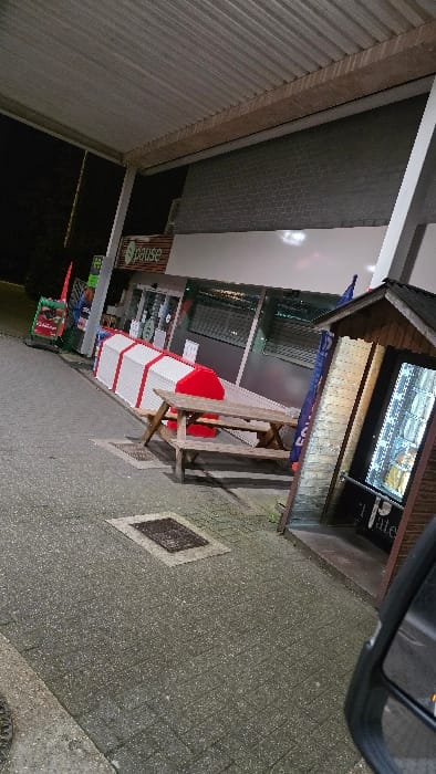 The scene depicts the exterior of a service station or shop at night, likely located in Herentals, Belgium. The area is illuminated by overhead lights from a corrugated metal canopy. The ground consists of grey paver stones with two visible metal utility covers.

To the left, a green and red advertising banner is visible, along with what appears to be a fuel dispenser structure. Further right, a building with a grey brick upper facade and large glass windows below is visible. A prominent green "pause" logo is affixed to the building's exterior above the entrance. Directly in front of the building, a long white structure with red striped accents is positioned, resembling a barrier or display unit.

In the center, a weathered wooden picnic table with attached benches is situated. A bright red object, possibly a cushion or cover, rests on the bench closer to the building. To the far right, a rustic, hut-like wooden structure houses a lighted vending machine displaying numerous drink cans. A partial white text, "Late," is visible on the bottom frame of the vending machine. A blue flag is also visible leaning against this wooden structure. The bottom right corner shows a portion of a black vehicle's side mirror. No individuals are present in the scene.
