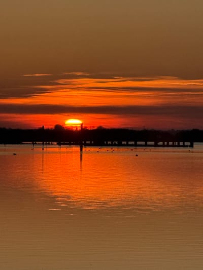 This image depicts a sunset over a body of water within Broadland, United Kingdom. The sun, a bright yellow-orange disc, is partially obscured by the distant horizon, positioned centrally near the bottom of the upper third of the frame. The sky above is dominated by stratified horizontal clouds, displaying intense shades of orange and red, transitioning to muted brownish-orange towards the top. A dark, silhouetted landmass forms the horizon line, featuring trees and indistinct structures, including a prominent cylindrical shape near the sun's position.
The midground is occupied by the calm water surface, which vividly reflects the vibrant orange and red hues of the setting sun and sky. Extending horizontally across the midground, partially submerged in the water, is a line of dark, uniform posts or pylons, possibly remnants of a pier or bridge, casting vertical reflections onto the water. Several small, dark avian figures are visible scattered across the water's surface. The foreground consists of the water, showing subtle ripples and a gradient of reflected light. No individuals or specific actions are discernible.
