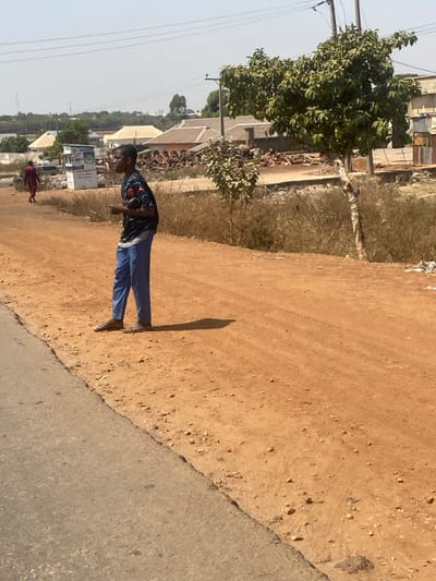 The scene depicts a sunny outdoor environment in the city of Lapai, Nigeria. In the foreground, a young male stands in profile, facing left, on a reddish-brown dirt path that borders a paved road on the left. He is wearing a dark, short-sleeved patterned shirt, loose blue trousers, and light-colored open-toed sandals. His hands are clasped in front of him, and he casts a distinct shadow to his right.
In the mid-ground, a larger green-leafed tree stands on the right, with a smaller similar tree to its left. Behind the main subject, another person in a long, dark red garment walks away from the viewer on a similar dirt path. The background features several light-colored, single-story buildings with varied roof styles, including what appears to be corrugated metal. To the right, behind the trees, piles of dark, irregular objects, possibly tires, are visible. Overhead utility lines traverse the clear, bright sky. A white signboard is partially visible in the distant left background. The overall environment appears dry and dusty with sparse, dry vegetation.