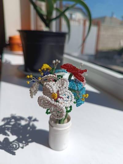 This is a close-up, eye-level, medium shot of a small, white vase holding a beaded flower bouquet. The bouquet consists of multiple flowers made from small, colorful beads, including white, red, blue, and yellow. Some of the flowers have green beaded stems and leaves. The vase is simple and white, with a textured surface. The bouquet and vase are placed on a white surface, possibly a windowsill, which is illuminated by sunlight casting a distinct shadow of the bouquet on the surface to the left. In the background, out of focus, is a black pot containing a green plant. Beyond the plant, a window shows a blurred view of buildings and a blue sky. The scene appears to be indoors, with natural light streaming in from the window. The image does not contain any individuals or any indication of the city of Nartan, Russia.