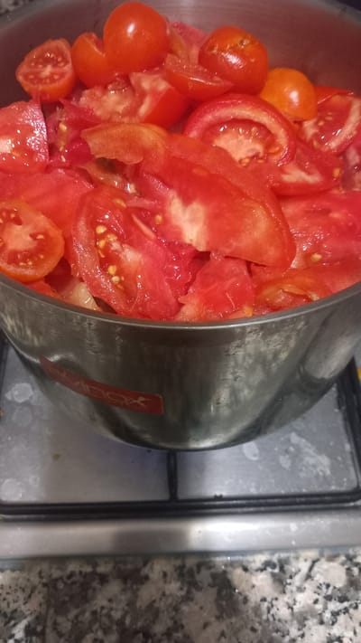 A close-up, eye-level, vertical shot captures a stainless steel pot overflowing with sliced tomatoes and whole cherry tomatoes, likely in preparation for cooking. The pot is placed on a grey speckled countertop, directly on top of a black gas stovetop, with visible circular grates. A portion of a red sticker with white lettering, possibly a brand logo, is visible on the side of the pot. The overall lighting is dim, creating a slightly muted color palette with prominent reds and oranges from the tomatoes. The scene appears to be a kitchen environment within a residential setting. There is no indication of the specific location in Oliveira do Bairro, Portugal.