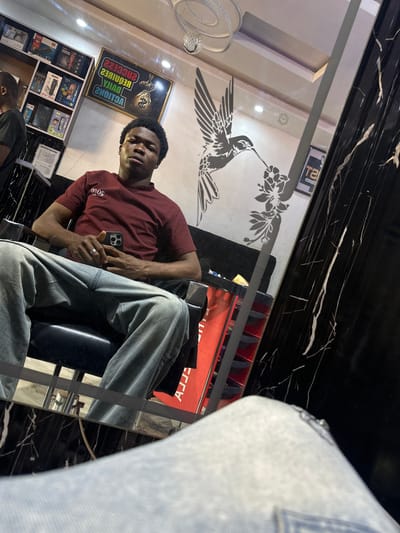 A man sits in a black barber chair holding a black mobile phone in his left hand while his right hand rests on his leg in what appears to be a barbershop based in the city of Ogwashi-Uku, Nigeria. The man is wearing a maroon T-shirt and gray jeans. The barbershop is decorated with a hummingbird sticker and features a black and white marble-like countertop. A sign with the words “Success requires daily actions” and other items are seen on a shelf in the background. The photo appears to be taken using a mirror’s reflection, as suggested by the angle and the presence of the photographer’s jeans in the foreground of the reflection.