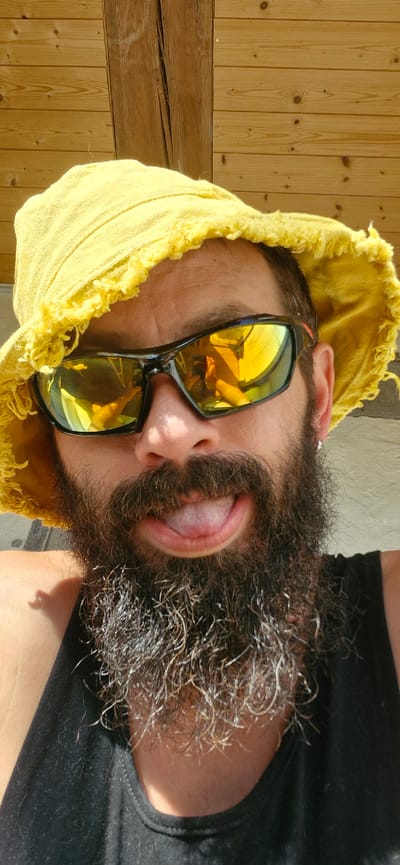 A selfie taken outdoors in bright sunlight features a person wearing a yellow bucket hat and reflective sunglasses. The person has a dark, full beard with some graying hairs, and their tongue is sticking out. The background consists of a wooden wall. The image appears to have been taken in Wattenwil, Switzerland.
