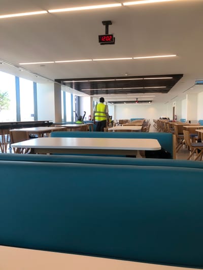 The image displays the interior of a large, brightly lit cafeteria or dining hall located in Hanak, Saudi Arabia. The ceiling is white, featuring multiple parallel linear LED light fixtures and a centrally suspended digital clock displaying "12:02" in red numerals. A dark, slatted ceiling panel runs down the center of the room. Large, floor-to-ceiling windows on the left wall provide natural light and an exterior view.
In the foreground, teal blue upholstered booth seating is visible, with a light-colored rectangular table featuring a white top positioned in front. The midground and background are occupied by numerous rows of dining tables, each with a light wood frame and a white top, accompanied by light wood chairs with blue upholstered seats.
A male individual is observed from the rear, walking towards the background, slightly right of the frame's center. He is dressed in a yellow high-visibility vest over a dark long-sleeved shirt and dark pants. At least two other individuals, also in high-visibility vests, are partially visible in the midground to the left, near the windows. A tall, dark piece of equipment, possibly a cleaning cart, stands near the windows on the left. A red fire extinguisher on a stand is present against a white wall in the midground. The dining hall appears largely unoccupied.