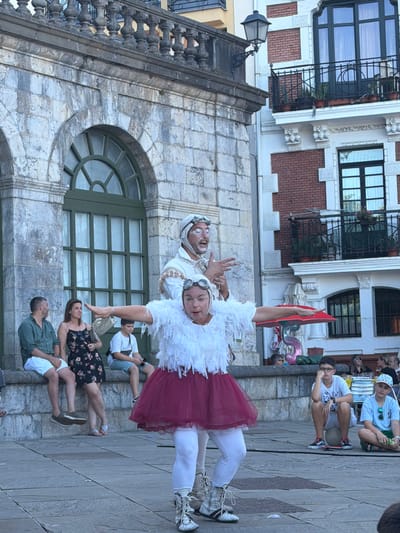 In Lekeitio, Spain, in front of a stone building with arched windows, two performers are engaged in a street performance. The performer in the foreground is wearing a white feathery costume, a burgundy tutu, white leggings, and combat boots. She is in the process of a dramatic pose, with her arms outstretched to the sides and her head tilted back. Behind her, another performer, dressed in a white outfit with goggles and white face paint, gestures animatedly with his hands. Several spectators are seated on a stone ledge in the background, watching the performance. To the right, a young boy wearing glasses sits on a ball, looking bored, while another boy in a blue shirt and hat watches the performance. The scene is set in a public square with cobblestone paving.