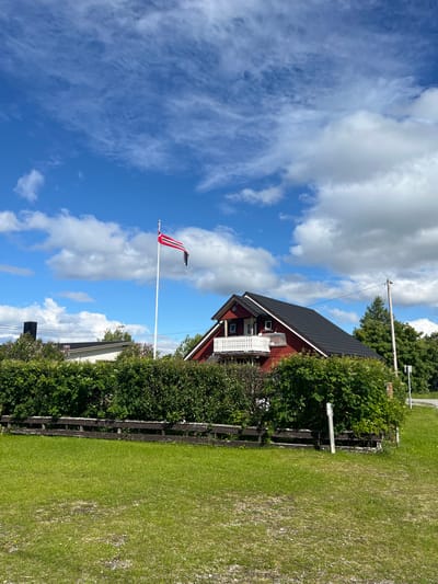 The image depicts a red wooden house with a dark roof and a white balcony in Lakselv, Norway. A flagpole with the Norwegian flag is prominently displayed in front of the house, against a backdrop of a blue sky with scattered white clouds. A dense green hedge runs along the front of the property, and a weathered wooden fence is visible in front of the hedge. To the left of the house, another building with a dark chimney is partially visible, and further to the left, a white building with a dark roof is also seen. The environment is suburban, with lush green grass in the foreground and trees in the background. No people are present in the image.
