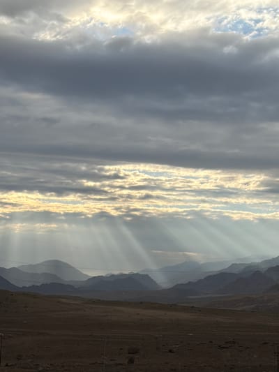 The image shows a panoramic view of a mountain range under a dramatic sky filled with clouds and sunbeams. The foreground is a desolate, dry landscape of brown earth with scattered rocks. In the middle ground, rolling hills and more distant mountains are silhouetted against the hazy sky. The sky is characterized by layers of dark and light clouds, with bright rays of sunlight piercing through them, creating a dramatic effect. There are no people or specific structures visible in the image, suggesting a remote, natural environment. The scene evokes a sense of vastness and natural beauty.