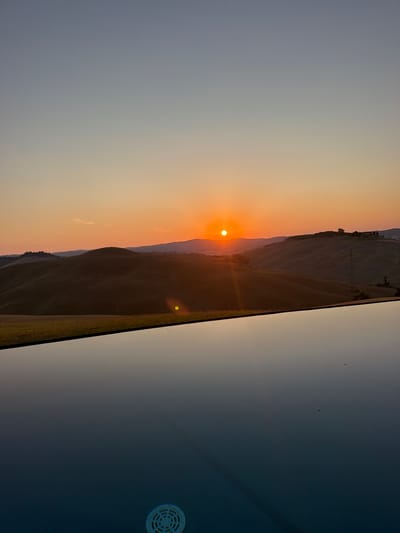 The image depicts a serene sunset over the rolling hills of Asciano, Italy. The sun, a bright orange orb, is setting on the horizon, casting a warm glow across the sky and landscape. The sky transitions from a pale orange near the horizon to a soft blue higher up. In the foreground, a calm, reflective swimming pool mirrors the colors of the sky and the surrounding hills. The water's surface is smooth, with subtle ripples and a visible drain cover at the bottom. The hills in the background are covered in golden-brown grass, characteristic of the Tuscan countryside. A few scattered trees and structures are visible on the hilltops, hinting at rural settlements. The overall scene conveys a sense of tranquility and natural beauty.