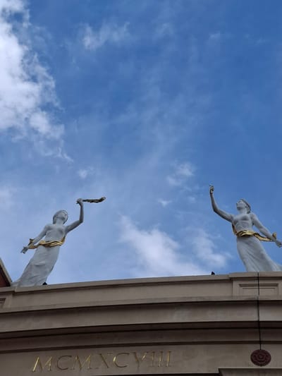 This is a low-angle, portrait-oriented photograph of two white statues of women against a bright blue sky with scattered white clouds. The statues are positioned on top of a building's cornice, which is decorated with the Roman numerals "MCMXCVIII" in gold lettering. Both statues are wearing golden sashes around their waists and appear to be holding objects. The statue on the left is holding a wreath or garland in its raised right hand. The statue on the right is holding a torch or flame in its raised right hand. A dark bird is perched on the cornice between the statues. The building's facade is light beige, with horizontal stone layers. The scene appears to be outdoors in daylight. This image was taken in Trafford, United Kingdom.