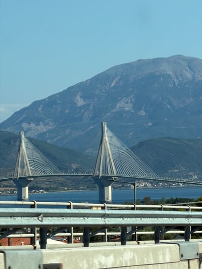 The image shows the Rio-Antirrio Bridge, a cable-stayed bridge spanning the Gulf of Corinth, connecting the Peloponnese peninsula to mainland Greece. The bridge is illuminated by daylight and stands against a backdrop of a clear blue sky and a large, hazy mountain range. The foreground features a concrete barrier and metal guardrails of a roadway, suggesting the photo was taken from a moving vehicle. Buildings and trees are visible on the shoreline in the middle ground. The bridge itself consists of two tall pylons supporting a vast network of cables, with a roadway passing through the center. The overall scene depicts a monumental piece of infrastructure integrated into a natural landscape.