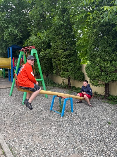 Two young boys are on a seesaw in a playground. One boy, wearing an orange long-sleeved shirt and a grey baseball cap, is on the left side of the seesaw, holding onto the red bar. He is looking up and to his left. The other boy, wearing a dark blue jacket and red shorts, is on the right side of the seesaw, also holding onto the bar. He is looking towards the camera with a smile. The seesaw has yellow and red metal supports and blue legs. The ground of the playground is covered in grey gravel. Lush green trees and foliage surround the playground, and a light-colored fence can be seen in the background. The lighting suggests it is daytime. No text is visible in the image. The scene appears to be a casual outdoor setting, likely a community or school playground.