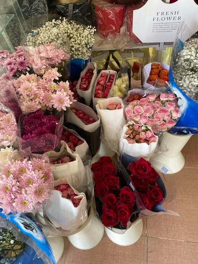 This media file captures an overhead view of a vibrant collection of flowers, likely displayed for sale. The subjects are predominantly bouquets of various flowers, including pink roses, red roses, orange roses, and pink and purple chrysanthemums. White baby's breath also features prominently. The flowers are arranged in a somewhat cluttered yet visually appealing manner, with some wrapped in clear plastic and others in decorative white paper or dark fabric.

The scene appears to be a flower shop or market stall. The setting is a flat surface, possibly a table or display, with a textured brown background suggesting a tiled or wooden floor. There are white bucket-like objects scattered amongst the flowers, and a blue plastic bag is visible in the upper left corner.

There are no people directly visible in the frame, but the presence of the flowers implies human activity related to their arrangement and sale. The time of day is not discernible, nor is the weather. The location context of Sidi Moussa, Algeria, is provided but not visually evident in the image itself.

Notable details include the text on a white card that reads "FRESH FLOWER" and a descriptive passage about the "flower Story of florist who loves flowers." Another visible detail is a QR code on one of the white bucket-like objects. The overall impression is one of abundant floral beauty.