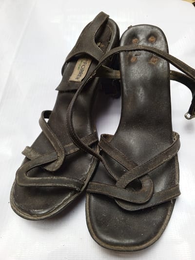 A pair of well-worn, dark brown leather sandals sit on a white surface. The sandals feature a strappy design across the toes, with a heel strap and a visible buckle. One shoe shows a label with the text "P. FORRER DESIGNS". The soles appear worn, and dust coats the leather straps. The sandals exude a vintage or retro aesthetic. Four metallic rivets can be seen on the top of the right heel.