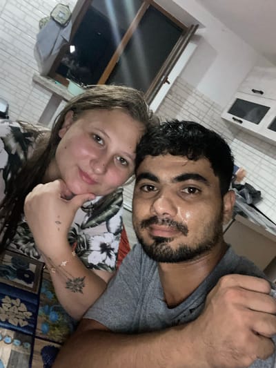 In Mitoc, Moldova, an indoor, dimly lit scene captures a close-up selfie of a couple. The woman, on the left, has light skin, brown hair, and is wearing a floral patterned shirt. Her left hand rests on her chin, and she has a tattoo on her left forearm with a bracelet. The man, on the right, has darker skin, short dark curly hair, a beard and mustache, and is wearing a gray t-shirt. He is holding the woman's arm with his right hand. They are positioned closely together, facing the camera. The background shows a kitchen area with white tiled walls and cabinets. A window is visible in the upper left corner, reflecting light.