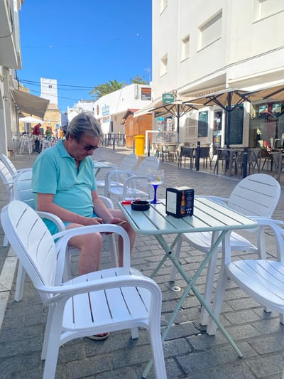 An outdoor cafe scene in Conil de la Frontera, Spain, features a middle-aged man sitting at a table, looking down at his phone. He is wearing a light blue polo shirt and blue shorts. In front of him on the table is a gin and tonic, a napkin dispenser with a beer advertisement, and an ashtray with a cigarette. The cafe appears to be on a street lined with white buildings, with awnings extending over the outdoor seating area. The ground is paved with cobblestones. In the background, other patrons are visible at tables, and some buildings have signs indicating "Taberna La Chanza" and "Delicatessen de Cadiz". The sky is clear and blue, suggesting a sunny day.