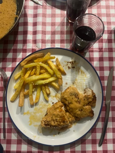 The scene depicts a dining table in Pedrouços, Portugal, covered by a red and white checkered tablecloth. A white circular plate with a blue rim is centrally positioned, containing a meal. On the left side of the plate, a quantity of golden-yellow french fries is arranged. On the right side, two pieces of chicken, coated in a thick, orange-hued creamy sauce, are visible; one piece is partially consumed, revealing white meat. Residue from the sauce and food particles are present on the plate, indicating ongoing consumption. To the left of the plate, a silver fork with a dark handle rests, and to the right, a silver knife is placed. In the upper right quadrant, two clear, ribbed drinking glasses contain a dark, reddish liquid, with one glass nearly full and the other approximately half-full. A black cooking pan, holding a substantial amount of the identical orange-hued creamy sauce, is partially visible in the upper left quadrant. The environment appears to be an indoor setting. No individuals are present in the image.