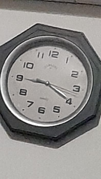 The image shows a clock mounted on a wall in Merlo, Argentina, located at coordinates -34.667 latitude, -58.733 longitude. The clock is octagonal in shape, with a black frame and a white face. The hands are black and the numbers are black on a white face. The time displayed on the clock is 3:50. The background of the image is a white wall.