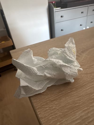 A crumpled white paper napkin lies on a light brown wooden table. The napkin has a textured surface with small, circular indentations. There is a faint yellow stain on one part of the napkin. In the background, a white cabinet with silver knobs is visible, along with a wooden shelf unit. The scene appears to be indoors.