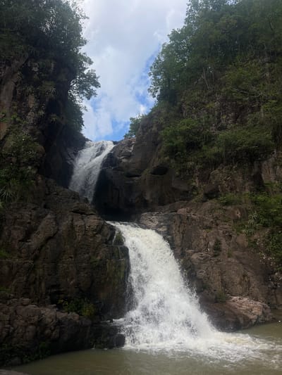 The image depicts a multi-tiered waterfall cascading down a rocky gorge in Arambala, El Salvador. Lush green vegetation, including trees and various plants, grows along the steep, dark rock faces that frame the waterfall. The water is white and frothy as it plunges into a calm pool at the bottom, which is a murky green color. The sky above is partly cloudy, with patches of blue visible between the white clouds. The overall scene conveys a natural, untouched environment. There are no people or animals visible in the image.