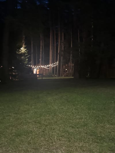 A nighttime outdoor scene shows a manicured lawn in the foreground, leading to a wooded area where string lights are hung between trees, illuminating a building in the background. The building appears to be a rustic cabin or lodge, with a warmly lit window. The trees are tall and slender, with dark foliage that blends into the night sky. The string lights create a festive and inviting atmosphere. The overall impression is one of a peaceful and serene evening in a natural setting. There are no people visible in the image. The location is described as being in the city of Kierwik, Poland.