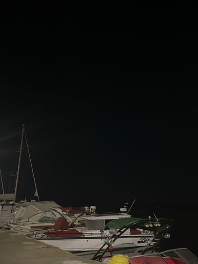 At night in Limenas Avdiron, Greece, several boats are docked at a marina. The foreground shows a concrete pier. A white and red motorboat with a green cover over part of its deck is prominently featured, with a yellow object on its deck. Behind it, another boat with a white hull and red seating is partially visible. Further back, a sailboat with tall masts stands out against the dark sky. The overall scene is illuminated by artificial lights, casting reflections on the water and highlighting the boats. The sky is dark and clear, with no visible celestial bodies. The water appears calm.