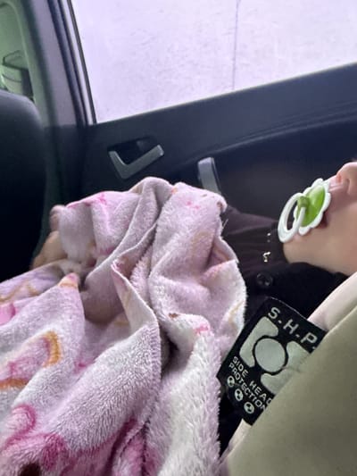 The image captures a close-up view of a baby or young child seated in what appears to be a car seat, viewed from inside a vehicle. The child's face is partially visible, showing their mouth with a green and white floral-shaped pacifier. They are wearing dark clothing, possibly a jacket or top.
A soft, pink and white patterned blanket, appearing to be plush or fleece, is draped over the child's lap and body, providing comfort. To the right of the image, a fabric tag is prominently visible, attached to what could be the car seat strap or headrest. The tag is black with white lettering and features the text "S.H.P." and "SIDE HEAD PROTECTION," along with an emblem that includes a circle and two small skull-like icons, indicating a safety feature. The car's interior is dark, with a light-colored headrest or trim visible above the tag.
Through the vehicle window on the left, an overcast or rainy sky is suggested by the diffused, light purple-grey light and subtle streaks on the glass, implying it might be daytime with poor weather. A dark car door frame or pillar is visible between the window and the child. Another dark handle or hook-like object is visible in the car interior near the window frame.
The overall scene suggests a child traveling comfortably and safely in a car, possibly in Kiselevka, Russia, given the provided location context.
