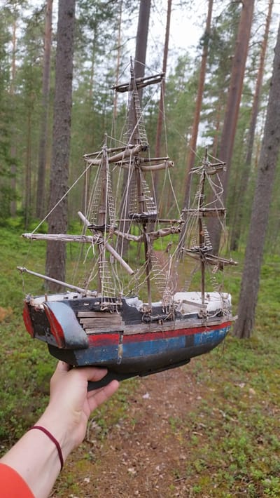 A hand wearing a maroon bracelet and an orange sleeve holds a weathered, wooden model ship with three masts and intricate rigging. The ship's hull is painted in alternating stripes of dark blue, red, and a lighter blue/grey. The ship is positioned in the foreground, with a blurred background of a forest. The trees are tall and slender with visible bark texture. The ground is covered with pine needles and sparse green vegetation. A dirt path is visible behind the ship. The overall lighting suggests an overcast or shaded environment. There is no visible indication of the city of Orekhovo, Russia.