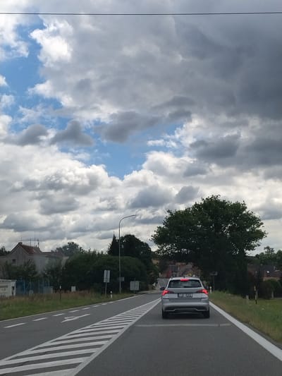 A silver Skoda Octavia is driving on a paved road in Bernartice, Czechia, on a cloudy day. The car has a Czech license plate that reads "7JO 8262". The road has white lane markings and a pedestrian crossing in the foreground. On the left side of the road, there are some buildings and trees. On the right side of the road, there is a grassy verge and trees. In the background, there are more buildings and trees. The sky is filled with dramatic clouds.