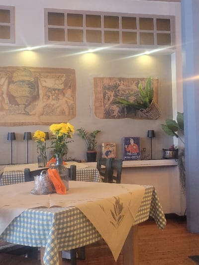 The scene depicts the interior of a dining establishment located in Λεχαινά, Greece. The foreground features a square wooden dining table covered with a blue and white checkered tablecloth, topped by a beige fabric overlay. This overlay has an embroidered olive branch design in its visible corner. On this table, a clear glass water bottle, a small clear glass holder containing what appears to be sugar packets or condiments, and a folded orange napkin are present. Dark wooden chairs are partially visible around this table and other adjacent tables.

In the mid-ground, additional dining tables with similar checkered tablecloths are arranged. One table to the left holds a clear glass vase with yellow chrysanthemum-like flowers. The background wall is painted a light neutral color, possibly beige or light grey, and features architectural details. Above a prominent white ledge, three downward-facing spotlights illuminate the wall. Below these lights, two large rectangular wall hangings or murals display classical-style imagery, including an ornate urn and figures resembling putti.

The white ledge supports several objects: multiple dark grey or black table lamps with conical shades, additional vases with yellow flowers, and various potted green plants. Also on the ledge are two vintage-style decorative metal signs: one depicting a man with the text "THIS GUY NEEDS A BEER!", and another depicting a woman with text that is partially obscured but appears to be "Hey mister, this one's on me!". A small, dark candlestick and a metal bowl containing purple grapes are also visible on the right side of the ledge. A woven basket holding a green plant hangs from the wall near the right mural. The upper portion of the wall includes a decorative recessed grid with multiple square sections. The floor consists of light brown wooden planks. No individuals are present in the image.