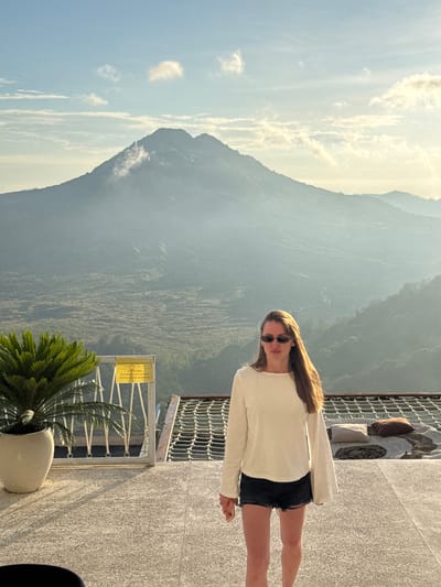 A long-haired woman wearing sunglasses, a light-colored long-sleeved shirt, and dark shorts stands on a concrete surface with a metal safety railing. Behind her is a mountainous landscape, possibly in the region of Kedisan, Indonesia. In the foreground, a potted plant sits near a sign with writing on it. A net-like structure is visible behind the woman. The sky is partly cloudy.