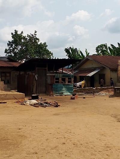 The scene is in Ogharefe, Nigeria, with coordinates 5.932 latitude and 5.671 longitude. There are two buildings in the scene: a larger, more modern building with a metal roof and a smaller, older building with a corrugated metal roof. The smaller building has a door on the side and a green metal wall. There is a black metal pot and some wooden beams in front of the smaller building. There are some clothes hanging on a line between the two buildings. There is a person standing next to the larger building. The ground is dirt and there are some leaves and debris on the ground. The sky is cloudy. There are a few trees in the background, including some banana palms.