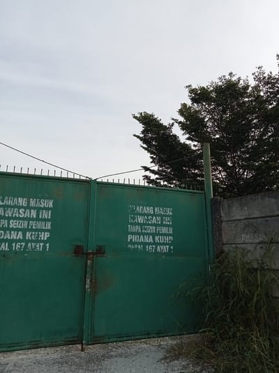 The image shows a closed, green metal gate with white lettering that reads "LARANG MASUK KAWASAN INI TANPA SEIZIN PEMILIK PIDANA KUHP PASAL 167 AYAT 1" on both the left and right sections of the gate. This translates to "NO ENTRY IN THIS AREA WITHOUT OWNER'S PERMISSION PENAL CODE ARTICLE 167 PARAGRAPH 1." The gate is secured with a rusty padlock and chain. Behind the gate, several trees with dark green foliage are visible against a cloudy sky. To the right of the gate, a concrete wall can be seen, and tall, dry grass grows in front of it and partially obscures the bottom right corner of the gate. The ground in front of the gate appears to be a gravel or asphalt surface. The overall scene depicts a restricted area with a warning sign, suggesting a private or guarded property. The location is identified as Teluk Kenidai, Indonesia.