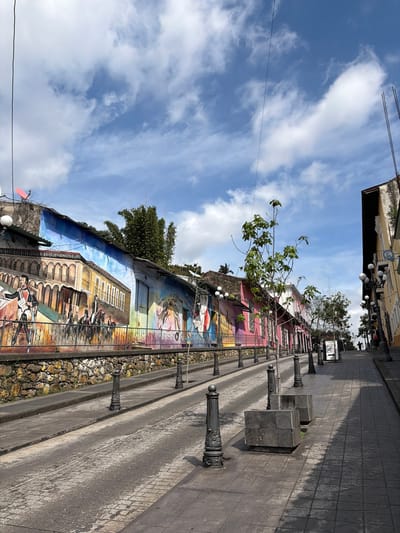 This image captures a vibrant, sunny street scene, likely a pedestrian walkway in Córdoba, Mexico. The perspective is angled slightly upwards, revealing a bright blue sky with scattered white clouds across the top. A thin utility wire stretches horizontally across the upper frame.
The dominant feature of the scene is a long, colorful mural painted on a low wall that runs along the left side of the street. The mural depicts diverse scenes, including figures in traditional attire, architectural elements like arched windows and balconies, and various activities, rendered in bright blues, reds, yellows, and greens. Behind and above this mural wall, parts of older buildings with a pale yellow facade and some greenery are visible. A prominent tree with bright green foliage extends into the mid-ground, appearing to grow from behind the mural. Further back, more dark green trees are visible.
To the right of the mural wall, a paved pathway or narrow street extends into the distance. This area is bordered by a series of dark, ornate bollards or decorative posts, each with a spherical top, spaced regularly along the length. Interspersed among these bollards are several large, dark, cubic blocks, possibly planters or seating. The paving itself appears to be a textured, dark grey material, possibly cobblestones or patterned concrete.
The clear blue sky and bright light suggest it is daytime, likely morning or early afternoon. The scene is static, with no discernible human activity or event taking place. The colorful mural and sunny weather evoke a cheerful and culturally rich atmosphere. No legible text is visible in the image.
