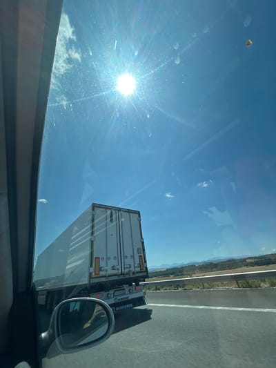 The image shows a view from inside a vehicle, looking out at a bright sunny day with a blue sky. The sun is high and appears very bright, causing lens flare. A large white semi-trailer truck with "KRONE" branding is visible in front and to the right, on a highway. The truck has various markings and lights on its rear. The vehicle's side mirror is in the lower left portion of the frame, reflecting a person's hand holding a phone. Beyond the highway, there is a landscape with fields and hills. There are some smudges or dust particles visible on the glass through which the picture is taken. The specific location in Vakarel, Bulgaria, is not discernible from the visual information provided.