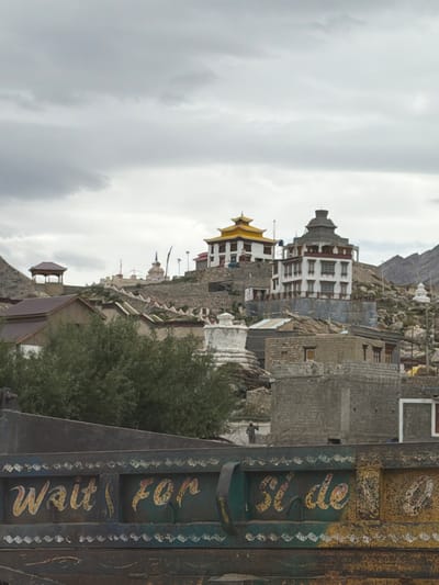 The image depicts a view of Kishrak, India, featuring a village nestled on a hillside with several buildings, some appearing to be monasteries or temples with distinct architectural styles, including white structures with red trim and golden roofs. The sky above is overcast with gray clouds. In the foreground, the back of a weathered, dark-colored truck is visible, adorned with colorful, hand-painted lettering. The text on the truck reads "Wait for Si de" in a stylized font with yellow and red accents, bordered by decorative white patterns. A few trees with green foliage are situated on the hillside, partially obscuring some of the buildings. The terrain is rocky and uneven, characteristic of a mountainous or hilly region. The overall impression is of a remote, possibly rural or historical, setting.