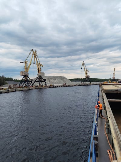 The image depicts a port scene in Shal'skii, Russia, under a cloudy sky. In the foreground, the edge of a large cargo ship is visible, with a person in an orange safety vest holding a thick rope. The ship's deck is dark and weathered.
Beyond the ship, a dark body of water stretches towards a concrete pier. Along the pier, several large yellow cranes are positioned. Two prominent cranes dominate the left side of the frame, standing tall with their booms extended. Behind them, a vast pile of gray aggregate material is stacked. Further down the pier, two more cranes are visible, smaller in scale.
The background features a dense line of green trees, suggesting a forested area bordering the port. The overall atmosphere is industrial and functional, with a focus on the logistics of cargo handling. The water is choppy, indicating a potentially windy or active marine environment.