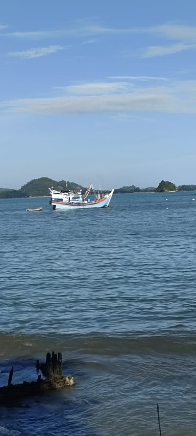 The image shows a serene coastal scene in Lhoktimon, Indonesia, under a partly cloudy sky. In the middle ground, a fishing boat with a blue hull and white superstructure is anchored in the calm, blue water. The boat has various fishing equipment visible, including nets and poles. To the left of the main boat, a smaller, lighter-colored dinghy is also present. The background features a verdant, tree-covered island with a small settlement visible at its base. Another smaller, rocky island is located further to the right. In the foreground, the dark, weathered remains of what appears to be a submerged structure or shipwreck are visible, with some wooden posts protruding from the water's surface, partially covered in green growth. The water in the foreground is slightly choppier than in the middle ground, with gentle waves breaking. The overall impression is one of tranquility and the daily life of a coastal community.