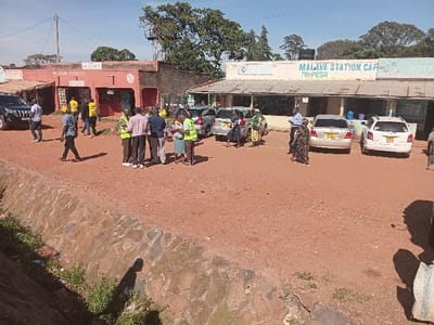The image depicts an unpaved commercial area in Butali Chegulo ward, Kenya, under a clear, bright sky.

In the foreground, a stone-lined drainage ditch, approximately 1.5 meters wide, runs horizontally across the frame, containing sparse green vegetation and visible litter. The primary ground surface beyond the ditch is reddish-brown dirt.

Approximately 20-25 individuals are present. A central cluster of 6-8 adults stands conversing; some in this group and other scattered individuals wear yellow or lime-green high-visibility vests. Other people are walking or standing, generally facing various directions.

On the left side of the frame, a two-story building with a reddish-orange facade houses multiple small storefronts at ground level. A dark-colored SUV, possibly a Toyota Land Cruiser Prado, is parked facing right near this building. Behind the building, a metal water tower supports a black cylindrical water tank.

On the right, a longer, single-story building with a light blue and white painted facade is visible. Its signage clearly reads "MALAVA STATION CAFE," and additional signs advertise "M-PESA" and "TOTAL." Several light-colored sedans and station wagons, identifiable as Toyota models, are parked in front of this building. Visible Kenyan license plates include KCA 481X, KCA 494X, and KCA 497X.

Trees are visible in the background and extending along the sides of the scene. Utility poles with overhead lines are also present. Shadows are distinct, indicating direct sunlight from a high angle.