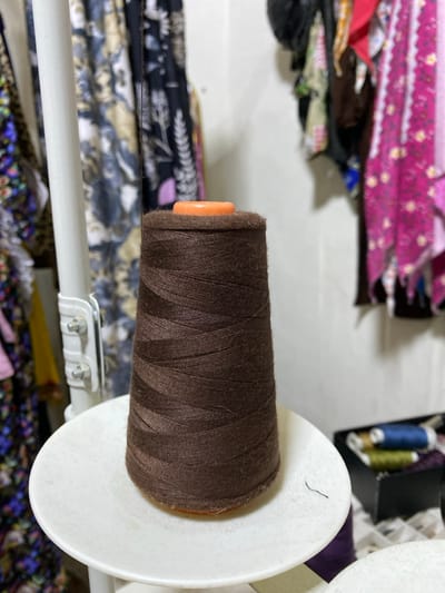 A large, conical spool of tightly wound dark brown thread, featuring an orange plastic core, rests centrally on a white circular plastic platform. A small, dark fiber lies on the platform to the right of the spool. To the left, a vertical white pole with metal fasteners is visible. The background on both sides is out of focus, revealing hanging textiles. On the left, patterned fabrics in shades of blue, beige, and dark brown are visible. On the right, an assortment of vibrantly colored fabrics, including pink, red, and blue, hangs. In the lower right background, a black container holds smaller spools of thread, including blue and olive green. The scene represents a textile or sewing workspace in Sidi Chahmi, Algeria. No individuals are depicted.
